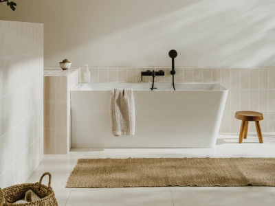 Minimalist bathroom featuring a white freestanding tub, beige tiled walls and floor, a towel over the tub, a wooden stool, and a woven basket.