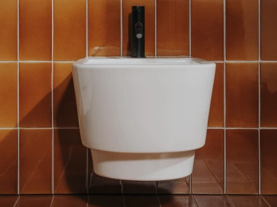 White wall-mounted urinal with a black flush valve, set against orange-brown tiled walls and floor.