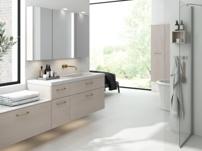 Bright modern bathroom with light-wood vanity, brass tap, large window view of greenery, wall cabinets, glass shower and towel rack.