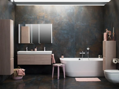 Modern bathroom with dark, metallic walls and a wall-mounted wooden double vanity with two sinks and a backlit mirrored cabinet; a white freestanding tub, pink towels and stool.