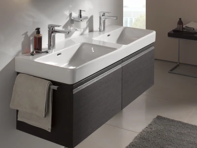 Double-basin white bathroom sink with two chrome taps on a dark wood vanity; soap dispenser, towel, grey rug and a side table with a bottle in the background.