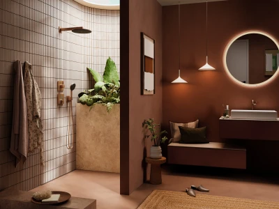 Modern bathroom with a left tiled shower and copper fixtures; right a brown vanity with a round backlit mirror, bench seating, plants and rug.