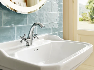 White porcelain bathroom sink with a chrome curved faucet, blue-green tiled wall, a round mirror, and a window showing greenery outside.