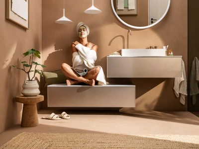 Person sits cross-legged on a floating vanity in a warm beige bathroom, wearing a face mask and white robe, with a plant and round mirror nearby.