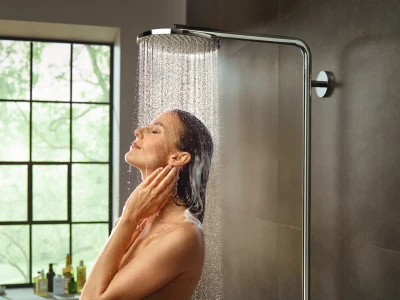 A person enjoys a rainfall shower in a modern bathroom, eyes closed, head tilted back, water streaming over wet hair, with bottles on the vanity and greenery outside.