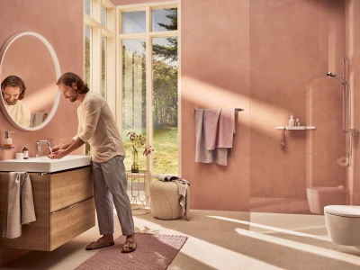 Person with long hair washes hands at a white sink in a pink bathroom, reflected in a round mirror with large windows opening to a garden.