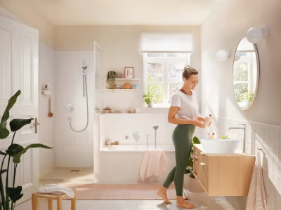 Bright bathroom with a person at a wooden vanity, holding a bottle at a white vessel sink; window light, round mirror, and plants around.