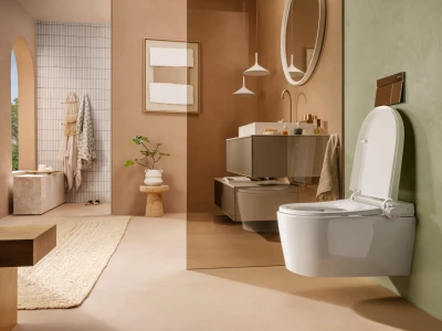A warm, modern bathroom with a wall-hung vanity, round mirror, pendant lights, a wall-mounted toilet, green wall, a plant on a stool, and a woven rug.