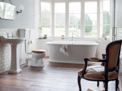 Bright vintage bathroom with a freestanding white bath by a bay window, pedestal sink, toilet with a wooden seat, and ornate dark wood armchair.