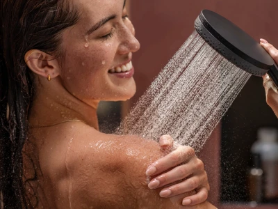Close-up of a person with wet hair smiling under a handheld shower, water cascading over their shoulder as they wash.