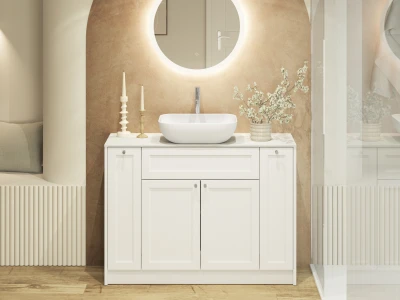 White bathroom vanity with round backlit mirror, vessel sink, and a vase of white blossoms on a beige wall; candles left, glass shower right.