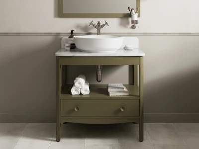Olive-green bathroom vanity with white marble top and round vessel sink, vintage tap, open shelf with towels and books, beige walls and tile floor.
