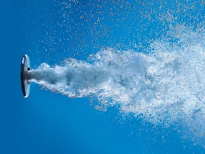 Water spurts from a circular nozzle on the left, forming a frothy, bubbly plume across a blue background.
