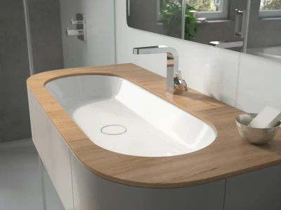 Modern bathroom vanity with an oval white sink set in a light wood countertop, chrome tap, white cabinetry, large mirror and a small bowl with soap.
