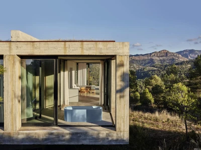 Modern concrete house with glass doors opens to a terrace; a blue circular tub sits outside while a seating area and distant mountains are visible.