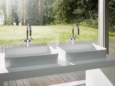 Two white rectangular sinks on a white countertop with chrome curved taps, in front of a large glass window overlooking a green garden and wooden deck.