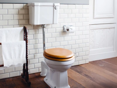 White toilet with a wooden seat in a vintage-style bathroom, white tiled walls, a towel rack with white towels, toilet paper, and wooden flooring.