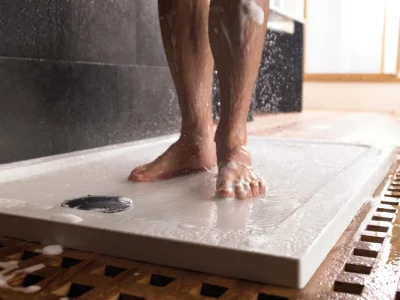 Bare feet standing on a white shower tray, water droplets spraying around with a drain visible to the left in a bathroom.