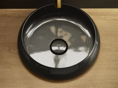 Top-down view of a round, black glossy vessel sink with a central drain, set into a light wood countertop; a gold stem rises above.