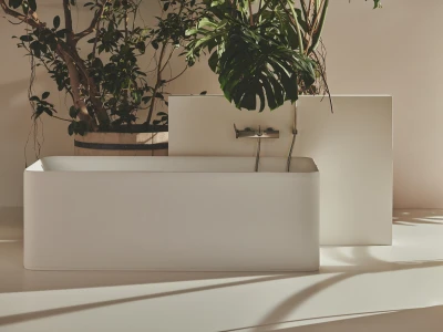 Minimalist bathroom with a white freestanding tub, white back panel and wall-mounted faucet, and a large leafy plant in a wooden pot; soft beige walls and shadows.