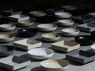 Neutral ceramic bowls and square dishes, both round and square, arranged on a dark floor in a geometric, shadowed display.