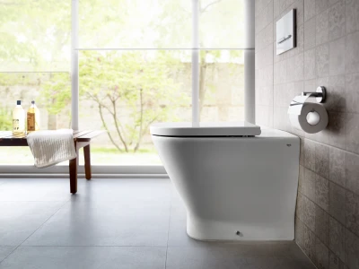 Modern bathroom with a sleek white toilet, a wooden bench with a folded towel and two soap bottles, and a large window showing greenery outside.