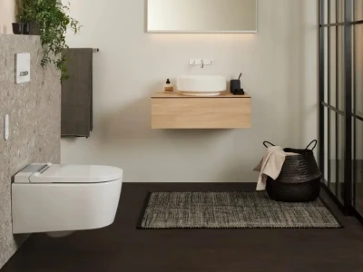 Modern bathroom with a wall-mounted wooden vanity, round sink, backlit mirror, toilet, plant, towel, rug and a black woven basket by glass partition.