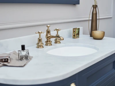 Gold brass faucet with two handles on a white marble bathroom sink, mounted in a blue vanity; a tall bronze vase and small bowl sit to the right.