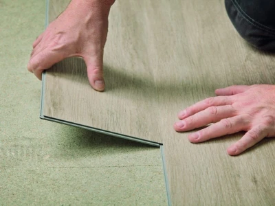 Two hands fit a wooden floor plank over green underlayment, aligning its edge during flooring installation.