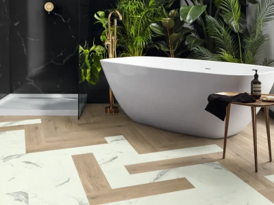 Modern bathroom with a white freestanding tub, gold tap, glass shower and lush tropical plants; light wood and marble geometric floor pattern.