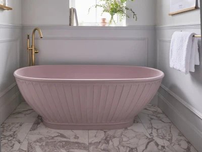 Powder pink freestanding tub with a brass faucet in a light grey bathroom, marble floor, plant on windowsill, and a white towel on a brass rail.