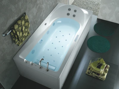 White corner whirlpool tub filled with pale blue water, chrome taps; grey walls, a green floral towel on a hook, two dark green mats, and a plant on towels.