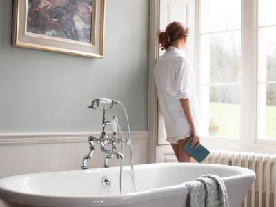 A person in a white shirt stands beside a white freestanding bathtub with a chrome faucet, holding a blue book and looking out a bright window.