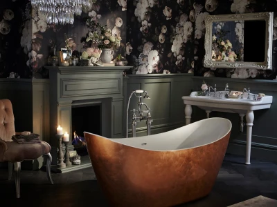 Luxurious bathroom featuring a copper freestanding tub, crystal chandelier, fireplace mantel with candles, floral wallpaper, an ornate mirror and a white sink.