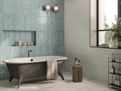 Modern bathroom with a freestanding grey tub against a teal vertical-tile wall, towel over the rim, recessed toiletries shelf, and a window with greenery outside.