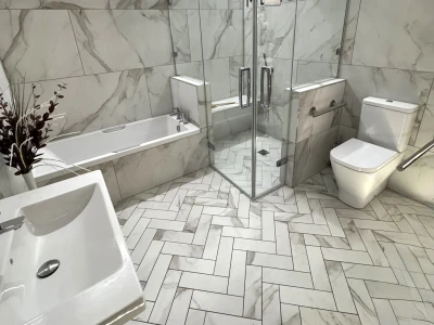 Modern marble bathroom with glass shower enclosure, toilet and sink; marble walls and a herringbone marble floor, with a plant by the sink.