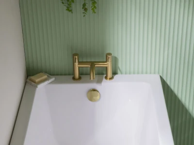 White freestanding bathtub with brass taps against a light green ribbed wall; soap on a small stone dish rests on the rim.