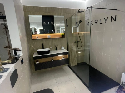 Modern bathroom showroom with a black toilet, white vessel sink on a black vanity, backlit mirror, wood paneling and a glass shower enclosure MERLYN.