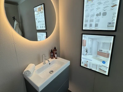 Small modern bathroom with a round backlit mirror above a white sink on a dark vanity, two brown bottles, a rolled towel, and two backlit product posters on the wall.
