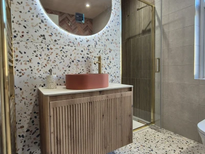 Modern bathroom featuring terrazzo walls and floor, round backlit mirror, pink circular basin on a wooden vanity, gold faucet, and glass shower.