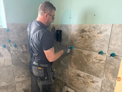 A construction worker tiles a bathroom wall with large stone-look tiles, wearing gloves and a harness, standing on a sheet with blue spacers.