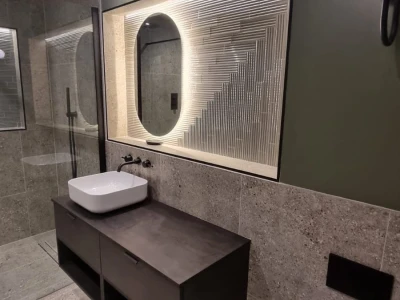 Modern bathroom with dark floating vanity and white vessel sink, backlit oval mirror in ribbed frame, glass shower enclosure, grey tiled walls and floor, and wall-hung toilet.