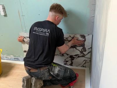 Person kneels on plywood floor, placing a marble-pattern tile along a pale green bathroom wall during renovation; wearing a black Inspira shirt.