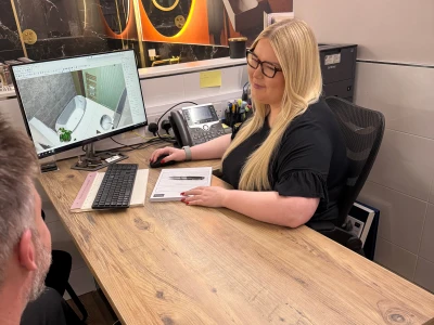 A person with long blonde hair and glasses sits at a desk, looking at a monitor displaying a 3D bathroom design; another person is visible opposite.