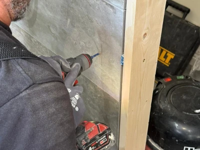 Person wearing safety glasses and gloves drills into a grey stone tile wall with a cordless drill; a wooden stud frame and red vacuum cleaner nearby.