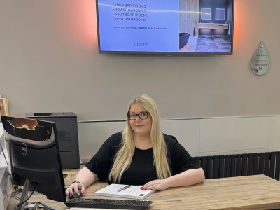 Person with long blonde hair and glasses sits at a wooden desk with a Dell monitor, keyboard and papers; a large wall screen shows a What We Can Do For You presentation.