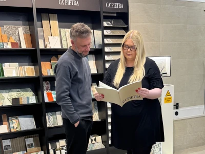 Two people in a tile showroom study a CA' PIETRA brochure; tall black display shelves behind them hold various tile samples.