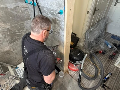 Person kneels on grey tiled bathroom floor beside a shower, drilling near a wooden frame; tools, a vacuum and hoses lie nearby.