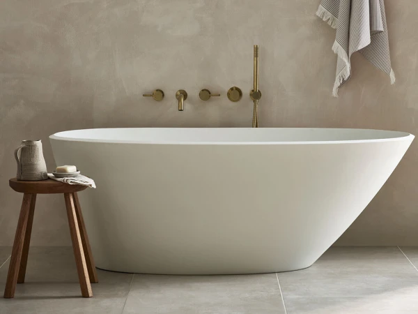 Minimalist bathroom featuring a white freestanding oval tub, brass wall fittings, beige textured walls, and a wooden stool with a jug and towel.