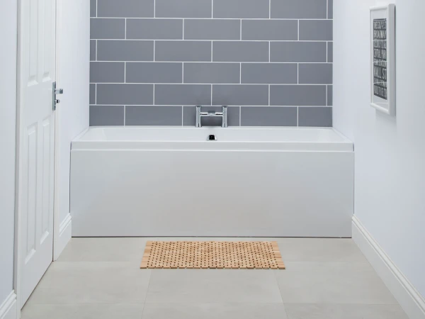 Bright bathroom with a white rectangular bathtub against a grey subway-tiled wall and chrome tap, wooden mat, door left and framed print right.